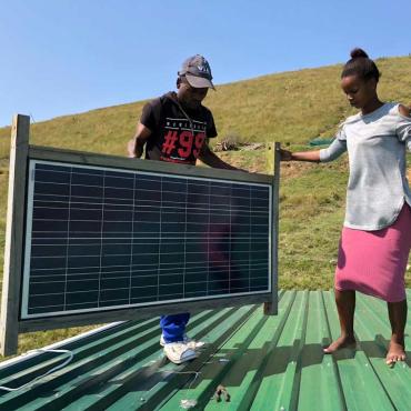 Image description: A man and a woman hold a solar panel on a roof