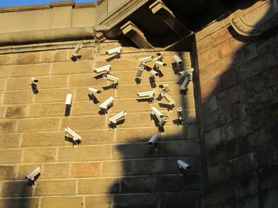 Image description: Numerous surveillance cameras on a wall 